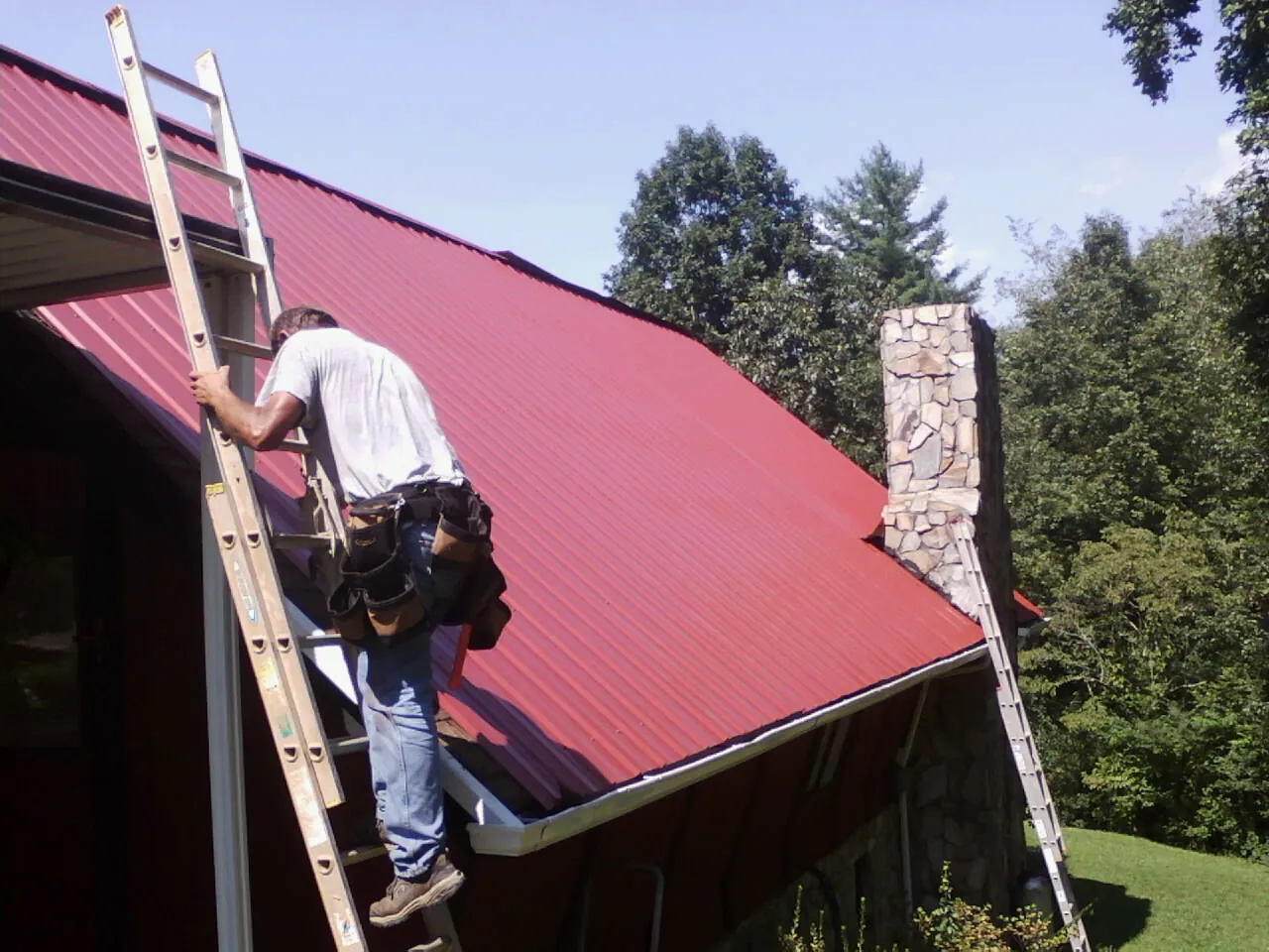 Roofer on a ladder beside a red metal roof and stone chimney — Soffit & Fascia Repair by Roofing Brothers in Vienna